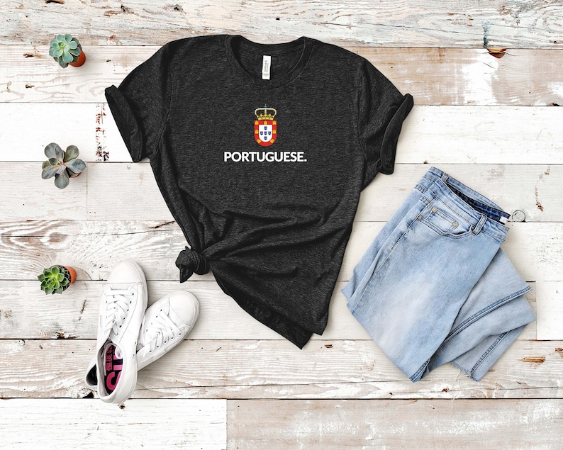 Może przedstawiać: Ciemnoszara koszulka z napisem "PORTUGUESE" i wzorem herbu. Koszulka jest połączona z jasnoniebieskimi dżinsami i białymi trampkami. Koszulka ma okrągły dekolt i kr&oacute;tkie rękawy.