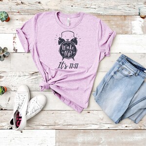 May include: A lavender t-shirt with a black alarm clock graphic and the text "Wake Up! It's 11:11". The shirt is laid out on a white wooden surface with a pair of white sneakers and blue jeans.