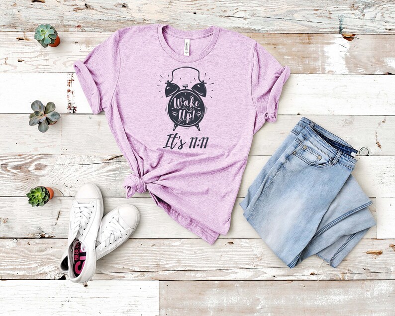 May include: A lavender t-shirt with a black alarm clock graphic and the text "Wake Up! It's 11:11". The shirt is laid out on a white wooden surface with a pair of white sneakers and blue jeans.