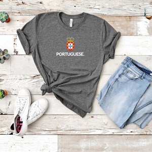 Może przedstawiać: Szara koszulka z napisem "PORTUGUESE" w kolorze białym, pod herbem. Koszulka jest połączona z jasnoniebieskimi dżinsami i białymi trampkami. Koszulka jest zawiązana w pasie.