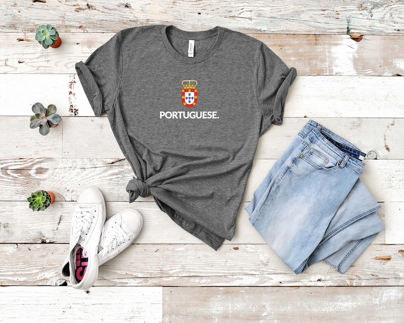 Może przedstawiać: Szara koszulka z napisem "PORTUGUESE" w kolorze białym, pod herbem. Koszulka jest połączona z jasnoniebieskimi dżinsami i białymi trampkami. Koszulka jest zawiązana w pasie.
