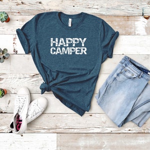 Puede incluir: Una camiseta azul turquesa con el texto "HAPPY CAMPER" en letras blancas desgastadas. La camiseta est&aacute; colocada sobre un fondo de madera blanca con un par de zapatillas blancas y unos vaqueros azules claros.