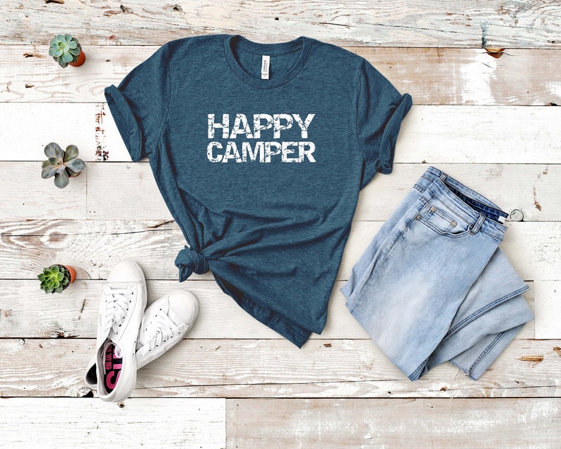 Puede incluir: Una camiseta azul turquesa con el texto "HAPPY CAMPER" en letras blancas desgastadas. La camiseta est&aacute; colocada sobre un fondo de madera blanca con un par de zapatillas blancas y unos vaqueros azules claros.