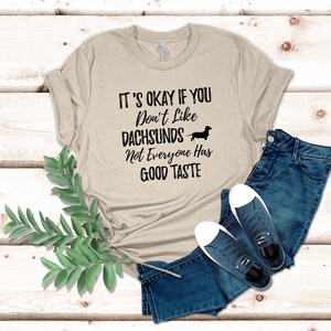 May include: A beige t-shirt with the text "It's okay if you don't like dachshunds. Not everyone has good taste." and a black silhouette of a dachshund.