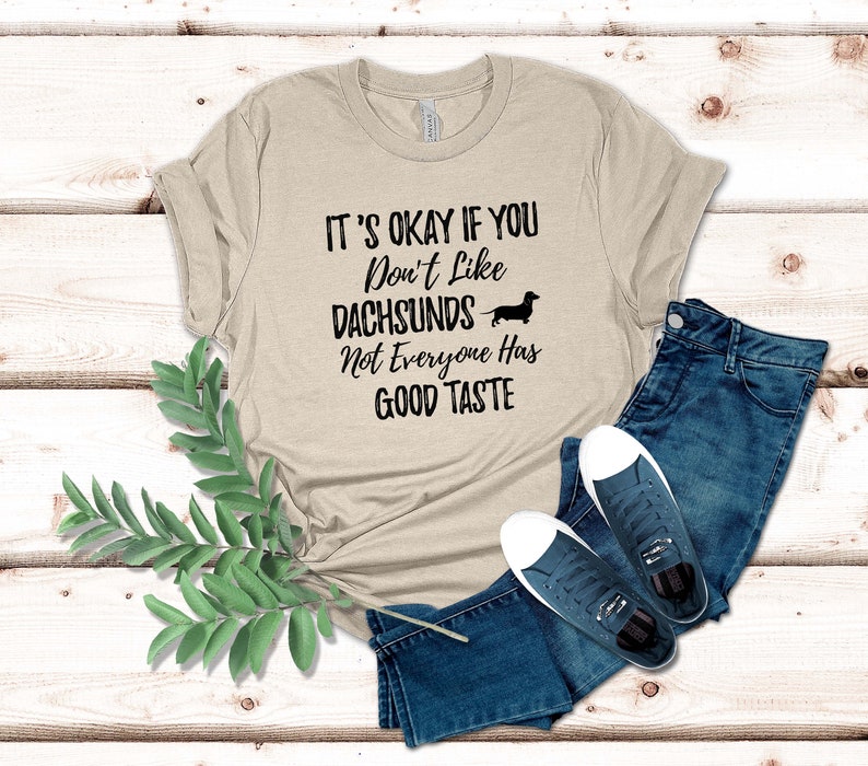 May include: A beige t-shirt with the text "It's okay if you don't like dachshunds. Not everyone has good taste." and a black silhouette of a dachshund.