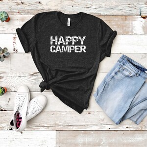 Puede incluir: Una camiseta negra con el texto "HAPPY CAMPER" en letras blancas. La camiseta est&aacute; colocada sobre una superficie de madera blanca con un par de zapatillas blancas y un par de pantalones vaqueros azules.