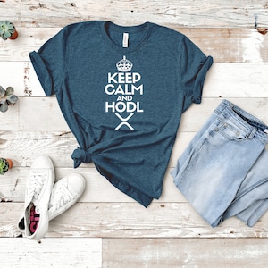Puede incluir: Una camiseta de color verde azulado con las palabras "KEEP CALM AND HODL" en blanco, un gr&aacute;fico de corona y un s&iacute;mbolo "X". Se combina con vaqueros azules de lavado claro y zapatillas blancas. Peque&ntilde;as suculentas en macetas est&aacute;n dispersas.