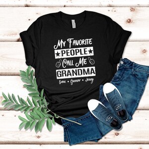 May include: Black t-shirt with white text that reads "My Favorite People Call Me Grandma" with the names Evan, Gunnar, and Jenny underneath.