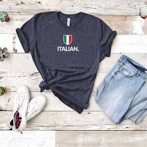 May include: A navy blue t-shirt with a white Italian flag graphic and the text "ITALIAN." printed on the front. The shirt is laid flat on a white wooden surface with a pair of white trainers and blue jeans.
