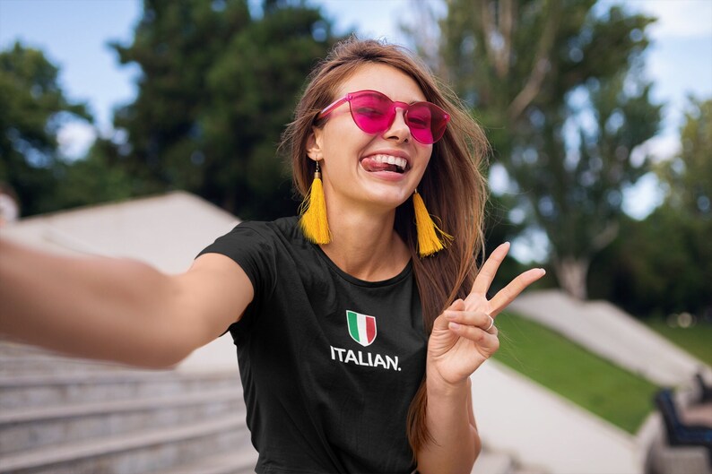 May include: A woman wearing a black t-shirt with a white Italian flag and the text "ITALIAN." She is wearing pink sunglasses and yellow tassel earrings. She is smiling and giving a peace sign.