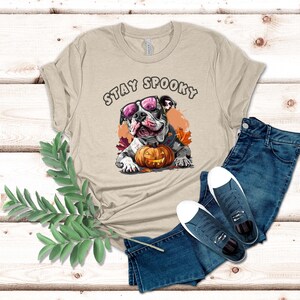 May include: A beige t-shirt with the text "Stay Spooky" printed on the front. A cartoon bulldog wearing sunglasses and a pumpkin is featured on the shirt.