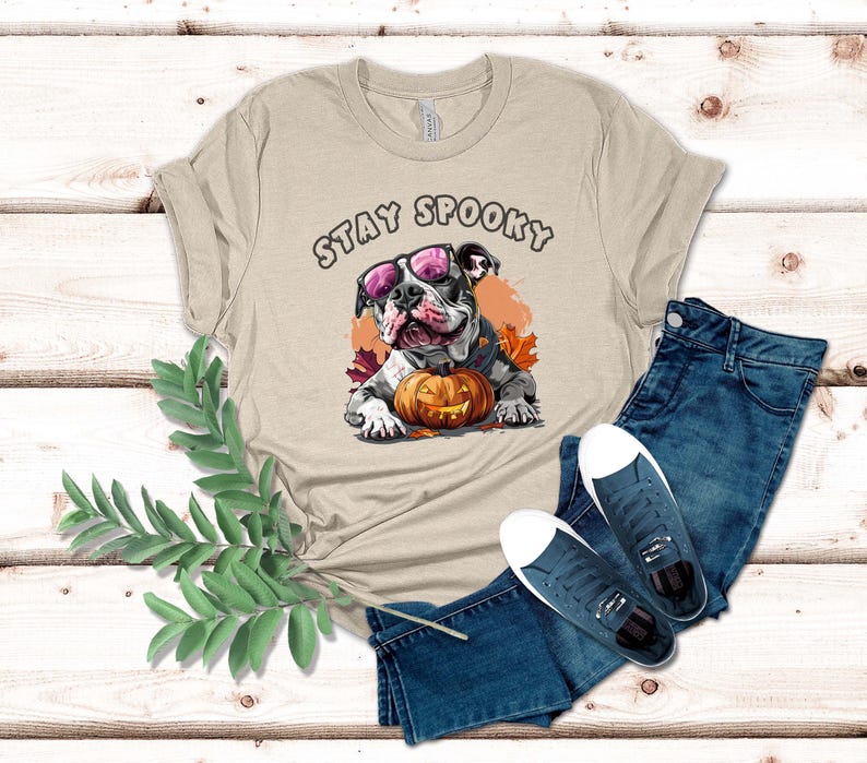 May include: A beige t-shirt with the text "Stay Spooky" printed on the front. A cartoon bulldog wearing sunglasses and a pumpkin is featured on the shirt.