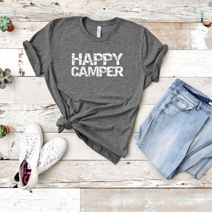 Puede incluir: Una camiseta gris con el texto "HAPPY CAMPER" en letras blancas desgastadas. La camiseta est&aacute; colocada sobre un fondo de madera blanca con un par de zapatillas blancas y unos vaqueros azules.