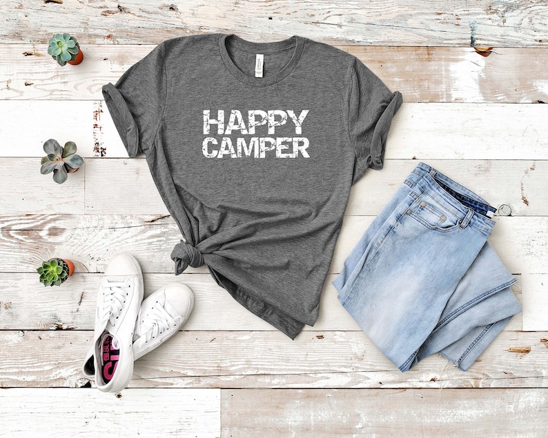 Puede incluir: Una camiseta gris con el texto "HAPPY CAMPER" en letras blancas desgastadas. La camiseta est&aacute; colocada sobre un fondo de madera blanca con un par de zapatillas blancas y unos vaqueros azules.