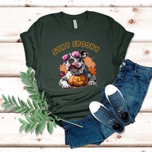 May include: A dark green t-shirt with the text "Stay Spooky" in orange letters. A cartoon bulldog wearing pink sunglasses is holding a jack-o-lantern pumpkin.