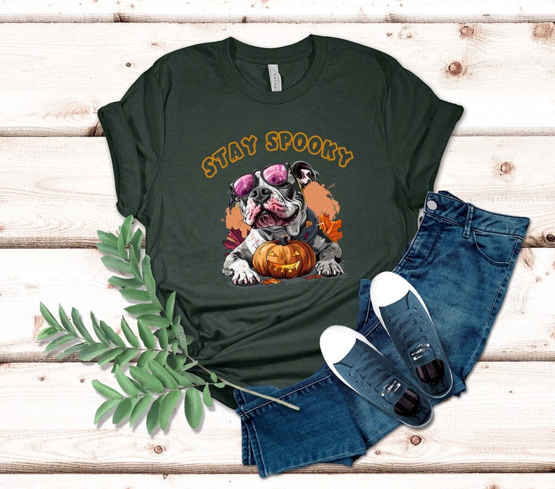 May include: A dark green t-shirt with the text "Stay Spooky" in orange letters. A cartoon bulldog wearing pink sunglasses is holding a jack-o-lantern pumpkin.