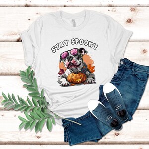 May include: White t-shirt with a graphic of a bulldog wearing pink sunglasses and holding a jack-o-lantern. The text "Stay Spooky" is printed above the bulldog.