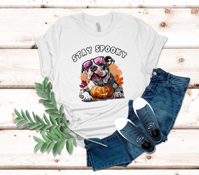 May include: White t-shirt with a graphic of a bulldog wearing pink sunglasses and holding a jack-o-lantern. The text "Stay Spooky" is printed above the bulldog.