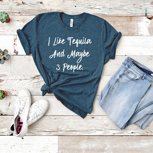 以下が含まれることがあります： 白地に「I like Tequila And Maybe 3 People.」と白で書かれたターコイズブルーのTシャツ。白い木製の表面に、白いスニーカー、薄いブルーのジーンズ、小さな多肉植物の鉢植えが3つ置かれている。
