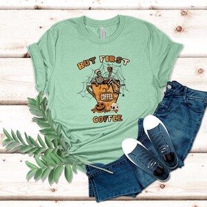 May include: A light green t-shirt featuring a graphic of two skeletons sitting in a coffee cup with the text "But First Coffee".
