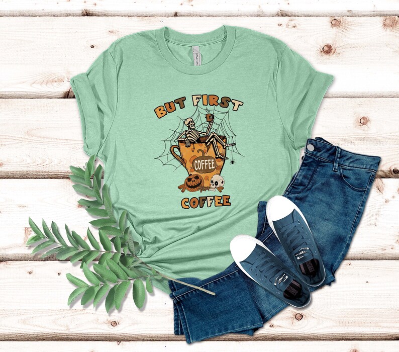 May include: A light green t-shirt featuring a graphic of two skeletons sitting in a coffee cup with the text "But First Coffee".