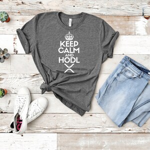 Puede incluir: Una camiseta gris jaspeado con el texto blanco "KEEP CALM AND HODL", una corona y un s&iacute;mbolo. La camiseta se combina con vaqueros azul claro y zapatillas blancas. Peque&ntilde;as suculentas en macetas est&aacute;n dispuestas alrededor de la ropa.