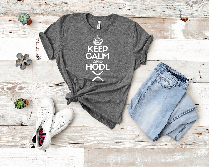 Puede incluir: Una camiseta gris jaspeado con el texto blanco "KEEP CALM AND HODL", una corona y un s&iacute;mbolo. La camiseta se combina con vaqueros azul claro y zapatillas blancas. Peque&ntilde;as suculentas en macetas est&aacute;n dispuestas alrededor de la ropa.