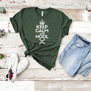 Puede incluir: Camiseta verde oscuro con el texto blanco &laquo;KEEP CALM AND HODL&raquo;, una corona y un s&iacute;mbolo &laquo;X&raquo;. La camiseta se combina con vaqueros azul claro y zapatillas blancas. El tema general est&aacute; relacionado con las criptomonedas.