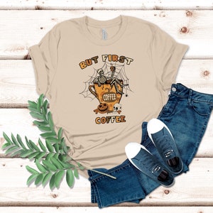 May include: A tan t-shirt featuring a graphic of a skeleton sitting in a coffee cup with a spider web and the text "But First Coffee".