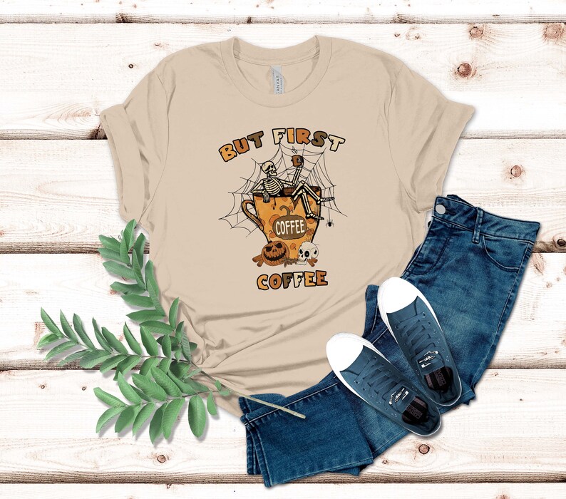 May include: A tan t-shirt featuring a graphic of a skeleton sitting in a coffee cup with a spider web and the text "But First Coffee".