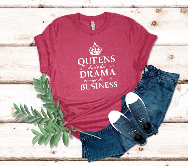 May include: A heathered dark pink short-sleeve t-shirt featuring a white graphic of a crown and the text "QUEENS don't do DRAMA we do BUSINESS" in a mix of serif and script fonts, paired with dark blue denim jeans and navy blue trainers.