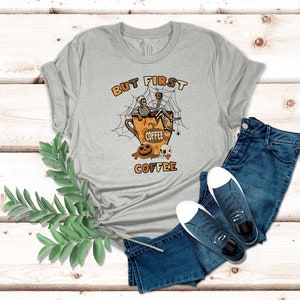 May include: A grey t-shirt with the text "But First Coffee" and a cartoon skeleton sitting in a coffee cup with a spider web and pumpkins.