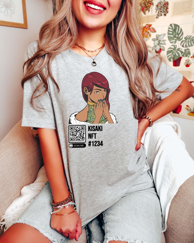 May include: A light gray t-shirt featuring a graphic of a woman with red hair and a gold collar. The text "KISAKI NFT #1234" is below the graphic, along with a QR code that says "SCAN ME".