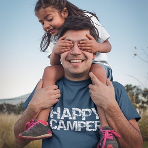 Puede incluir: Un hombre lleva una camiseta azul marino con el texto "HAPPY CAMPER" en blanco. Una ni&ntilde;a est&aacute; sentada sobre sus hombros y le tapa los ojos con las manos. Ambos est&aacute;n sonriendo.