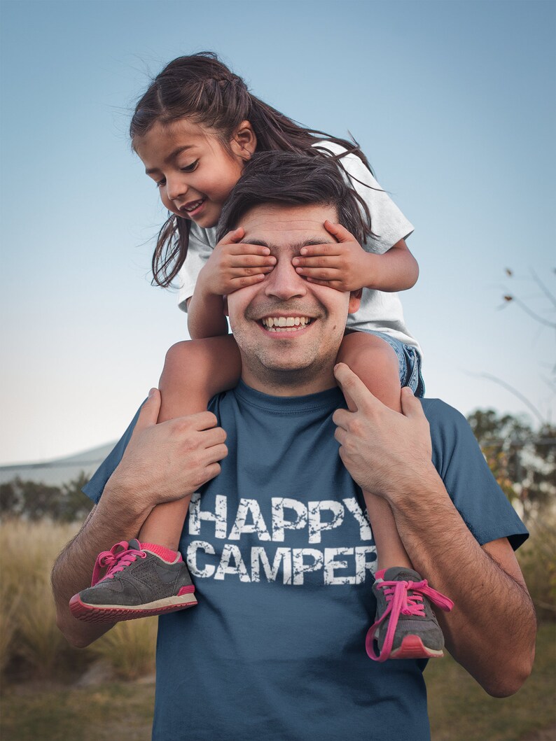 Puede incluir: Un hombre lleva una camiseta azul marino con el texto "HAPPY CAMPER" en blanco. Una ni&ntilde;a est&aacute; sentada sobre sus hombros y le tapa los ojos con las manos. Ambos est&aacute;n sonriendo.