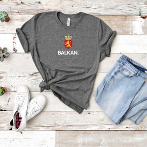 K&ouml;nnte beinhalten: Ein dunkelgraues T-Shirt mit einer wei&szlig;en Grafik eines L&ouml;wen mit einer Krone auf einem roten Schild und dem darunter gedruckten Wort "BALKAN."