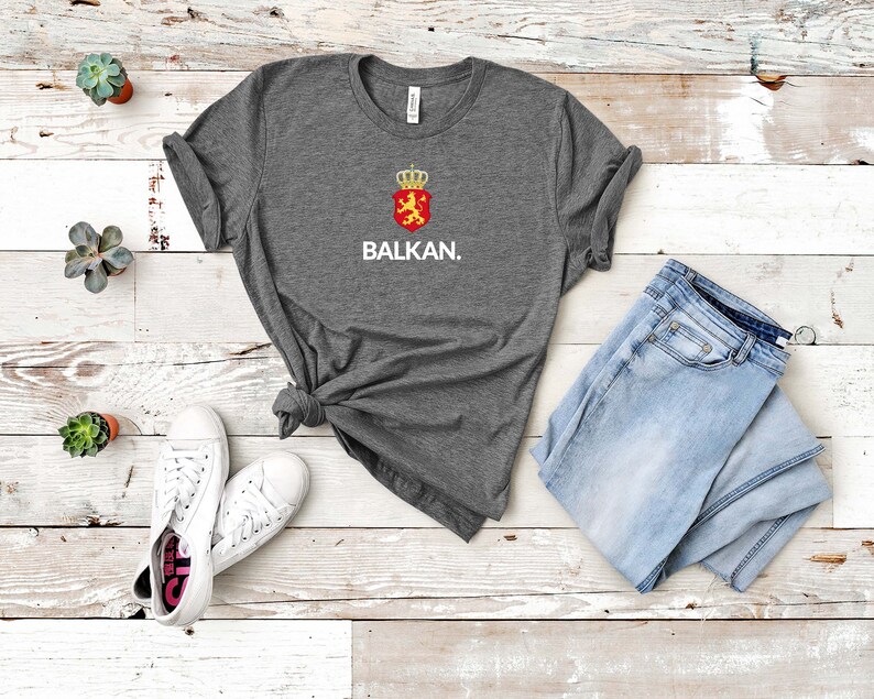 K&ouml;nnte beinhalten: Ein dunkelgraues T-Shirt mit einer wei&szlig;en Grafik eines L&ouml;wen mit einer Krone auf einem roten Schild und dem darunter gedruckten Wort "BALKAN."
