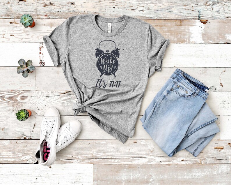 May include: Gray t-shirt with a black and white alarm clock graphic and the text "Wake Up! It's 11:11". The shirt is laid out on a white wooden surface with a pair of white sneakers and blue jeans.