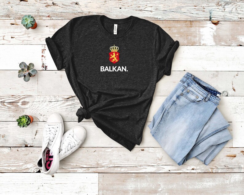K&ouml;nnte beinhalten: Ein dunkelgraues T-Shirt mit einer wei&szlig;en Grafik eines L&ouml;wen, der einen Schild mit einer Krone h&auml;lt. Der Text "BALKAN." ist unter der Grafik gedruckt.