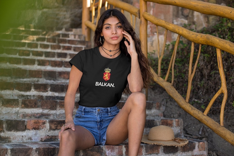 K&ouml;nnte beinhalten: Ein schwarzes T-Shirt mit einer wei&szlig;en Grafik eines roten Schildes mit einer goldenen Krone und einem L&ouml;wen sowie dem Text "BALKAN." Das Model tr&auml;gt das Shirt und eine blaue Jeans-Shorts.