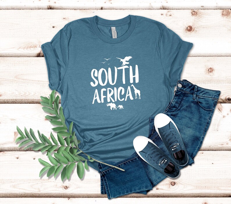 May include: A teal t-shirt with the words "SOUTH AFRICA" in white, along with silhouettes of an eagle, birds, a giraffe, and elephants. The shirt is paired with blue jeans and white-soled sneakers.