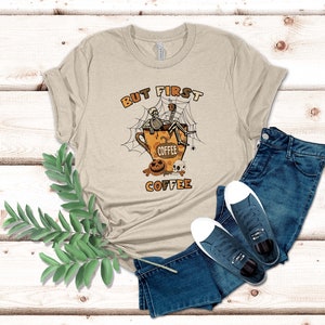 May include: A beige t-shirt featuring a graphic of a skeleton sitting in a coffee cup with a spider web and the text "But First Coffee".