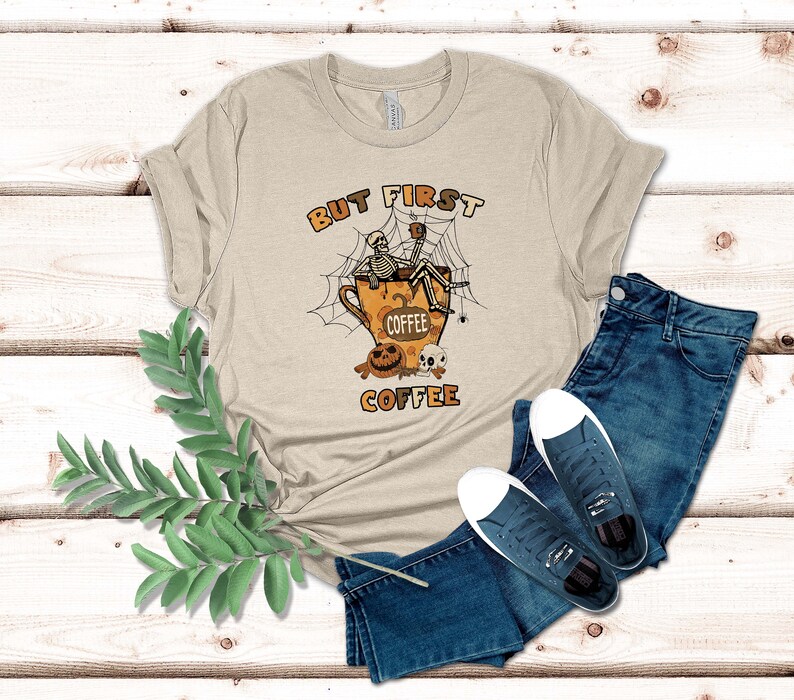 May include: A beige t-shirt featuring a graphic of a skeleton sitting in a coffee cup with a spider web and the text "But First Coffee".