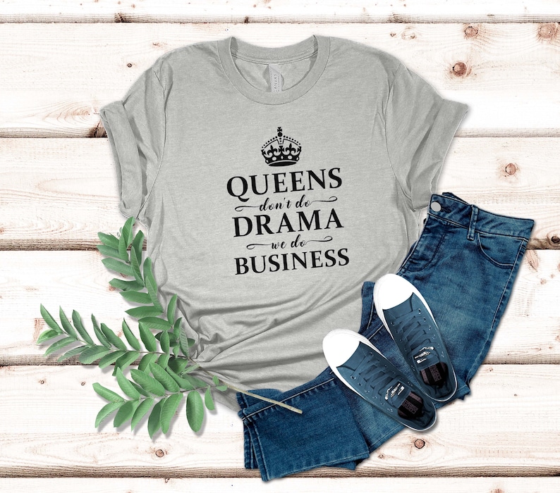 May include: A heather grey short-sleeved t-shirt featuring a black crown graphic and the text QUEENS don't do DRAMA we do BUSINESS, paired with dark blue denim jeans and a pair of dark blue canvas trainers with white laces and rubber soles.