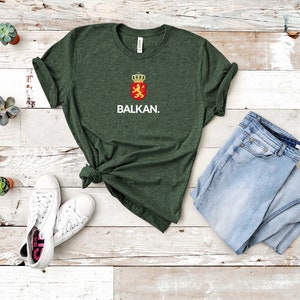 K&ouml;nnte beinhalten: Ein dunkelgr&uuml;nes T-Shirt mit einer wei&szlig;en Grafik eines L&ouml;wen mit einer Krone und dem Text "BALKAN." Das T-Shirt liegt auf einer wei&szlig;en Holzoberfl&auml;che mit einem Paar wei&szlig;en Sneakers und einer hellblauen Jeans.