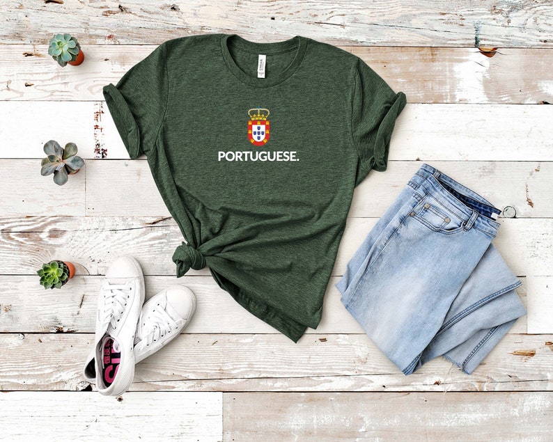 Może przedstawiać: Ciemnozielona koszulka z napisem "PORTUGUESE" w kolorze białym, pod herbem z koroną i tarczą. Koszulka jest połączona z jasnoniebieskimi dżinsami i białymi trampkami. Koszulka jest zawiązana w pasie.