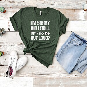 May include: Dark green t-shirt with the text "I'M SORRY DID I ROLL MY EYES OUT LOUD?" in white. The shirt is paired with light blue jeans and white sneakers. The shirt is knotted at the waist.