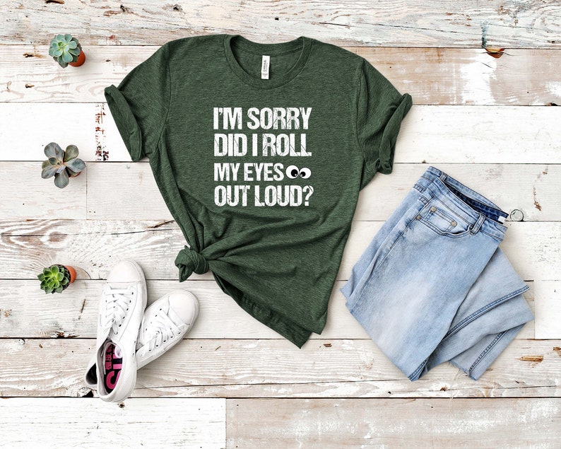 May include: Dark green t-shirt with the text "I'M SORRY DID I ROLL MY EYES OUT LOUD?" in white. The shirt is paired with light blue jeans and white sneakers. The shirt is knotted at the waist.