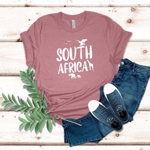 May include: A dusty rose t-shirt with the words "SOUTH AFRICA" in white, along with silhouettes of birds, a giraffe, and elephants. The shirt is paired with blue jeans and navy sneakers with white soles.
