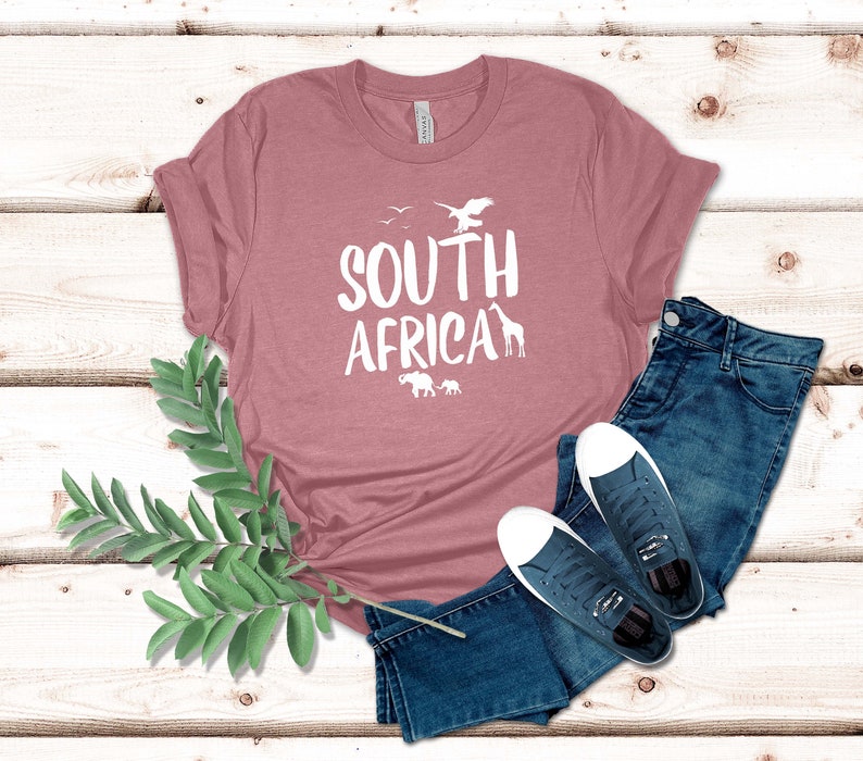 May include: A dusty rose t-shirt with the words "SOUTH AFRICA" in white, along with silhouettes of birds, a giraffe, and elephants. The shirt is paired with blue jeans and navy sneakers with white soles.